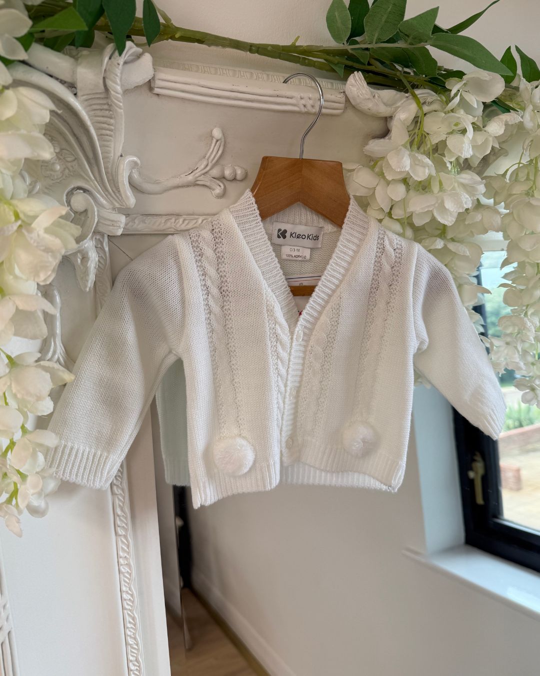 White knitted baby cardigan with cable knit detailing and pom pom trims.