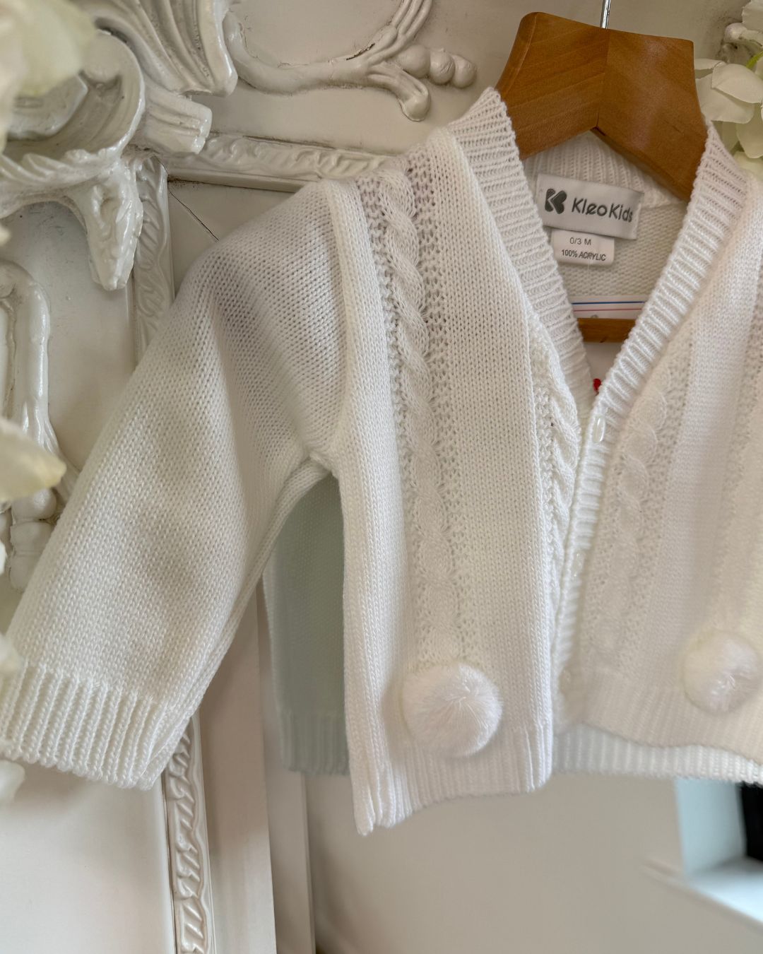White knitted baby cardigan with cable knit detailing and pom pom trims.