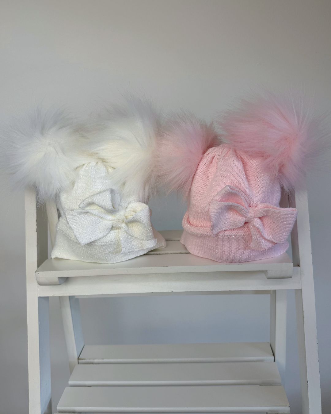 Shop the Little Nosh Sparkle Double Pom Hat, a cosy girls’ winter hat in white or pink with twin faux fur pom poms, a statement bow and warm ear covers.