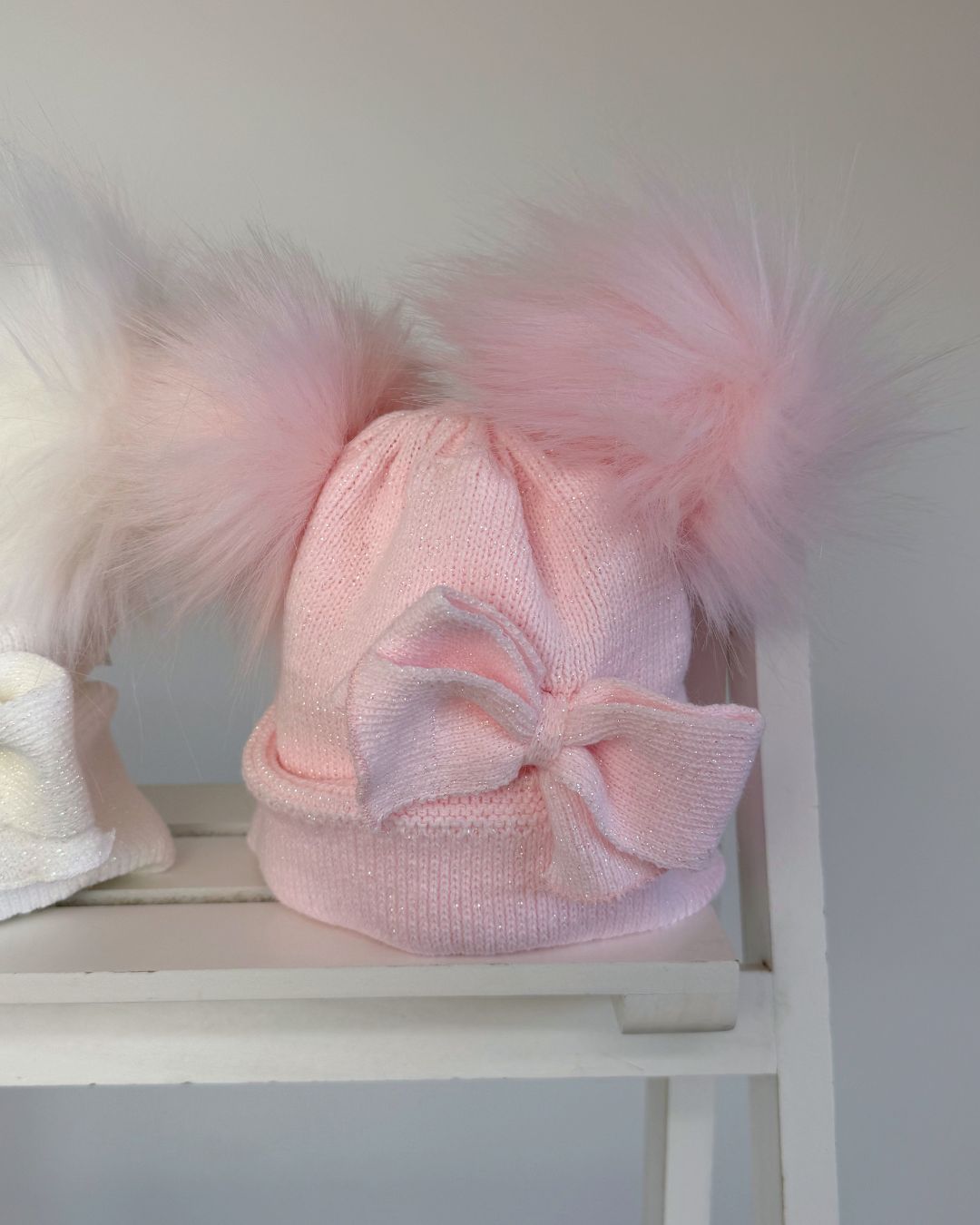 Shop the Little Nosh Sparkle Double Pom Hat, a cosy girls’ winter hat in white or pink with twin faux fur pom poms, a statement bow and warm ear covers.