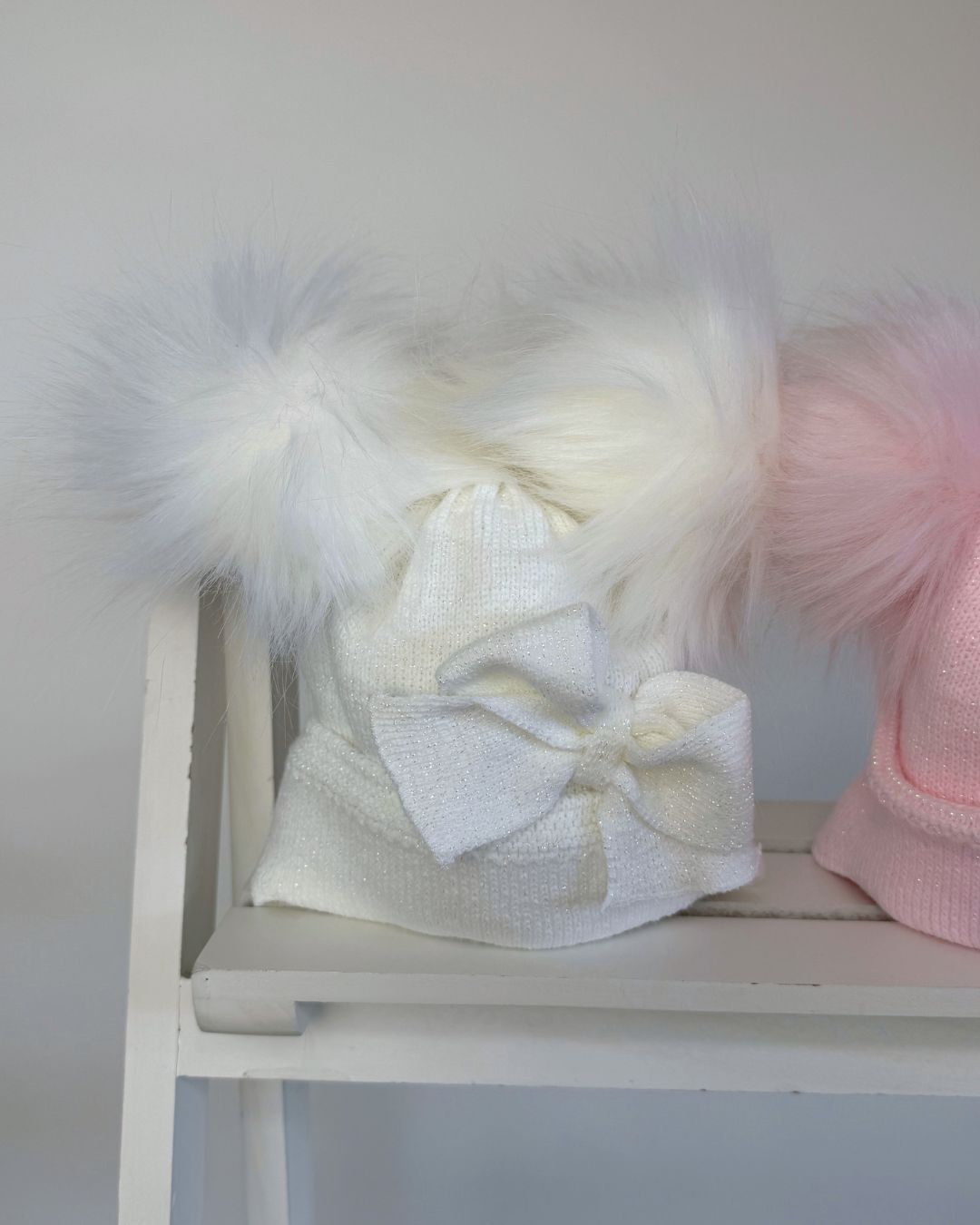 Shop the Little Nosh Sparkle Double Pom Hat, a cosy girls’ winter hat in white or pink with twin faux fur pom poms, a statement bow and warm ear covers.