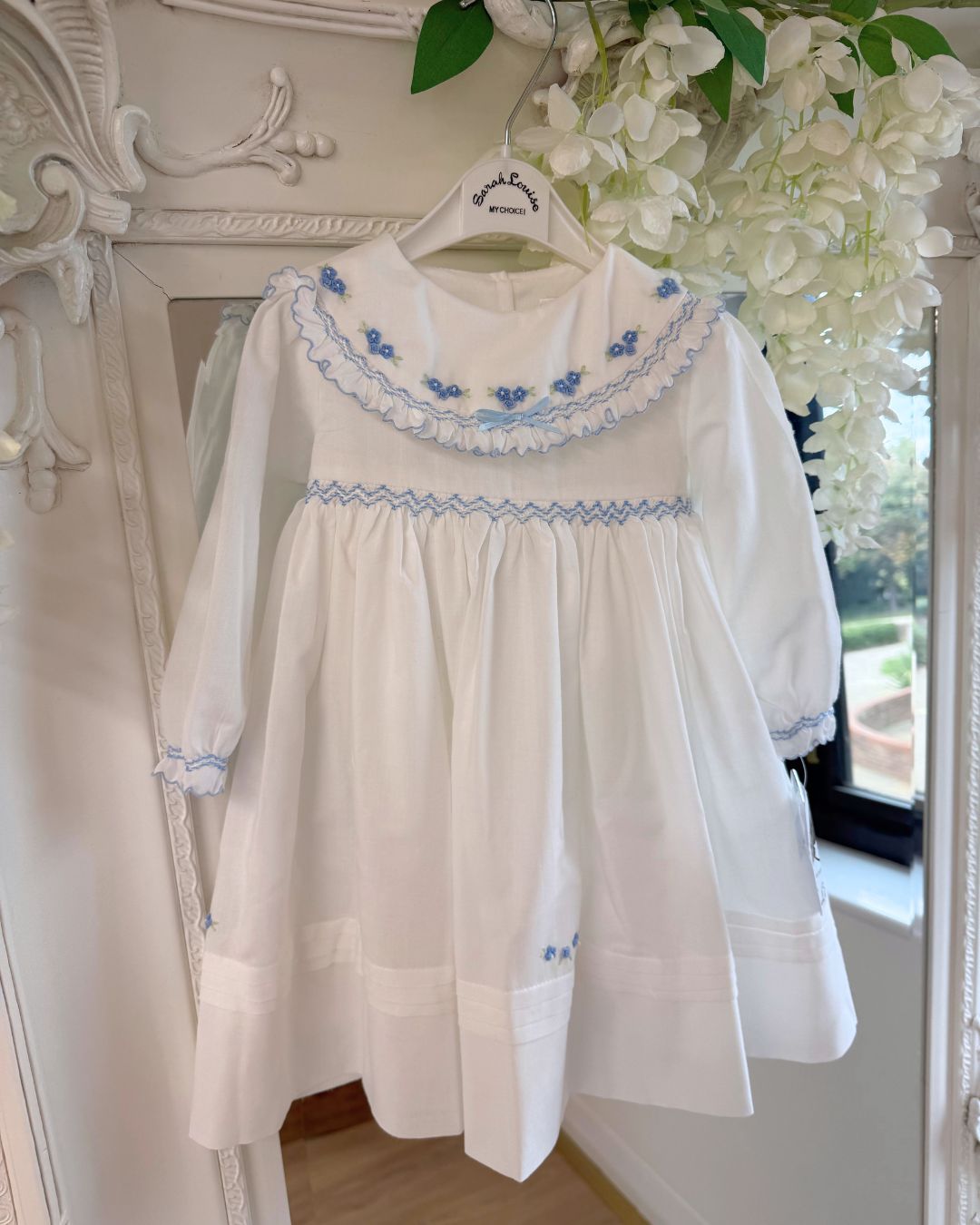 Sarah Louise Eleanor ivory girls smocked dress with blue floral embroidery, frilled collar, long sleeves and bow back tie.
