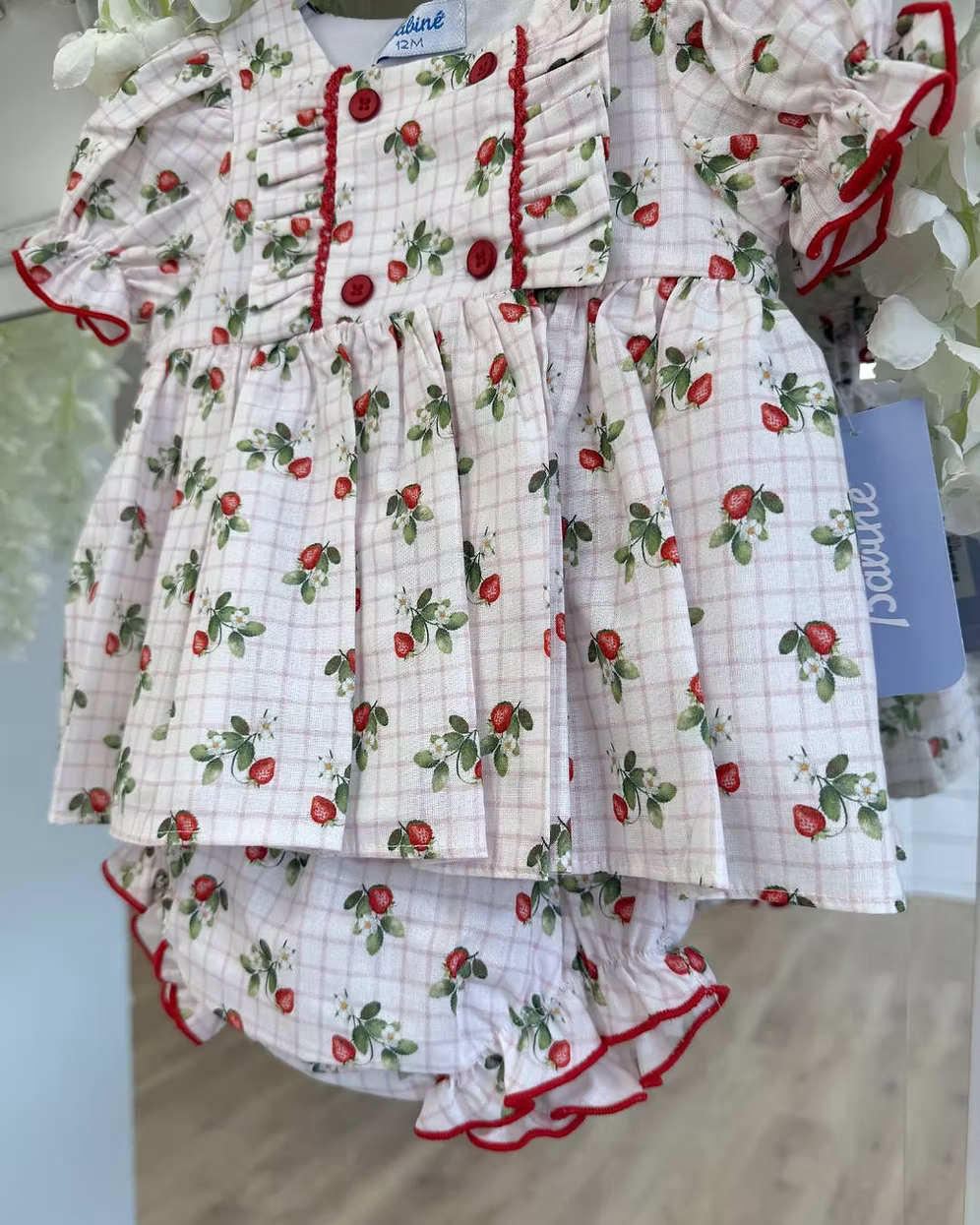 Off-white and pink check baby dress with strawberry print, frill chest detail, puff sleeves, red stitch trims, matching pants, and bow back fastening.