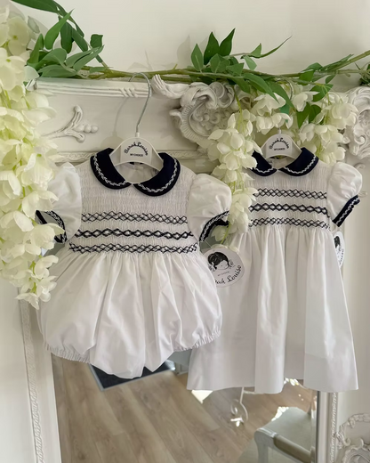 Baby girls white bubble romper with navy Peter Pan collar, sleeve trim and full smocked chest. Along with the matching sibling dress.
