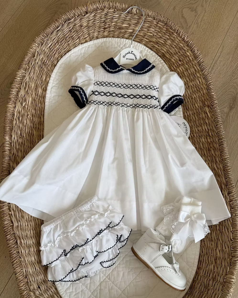 White baby dress with navy details and white shoes on a woven basket