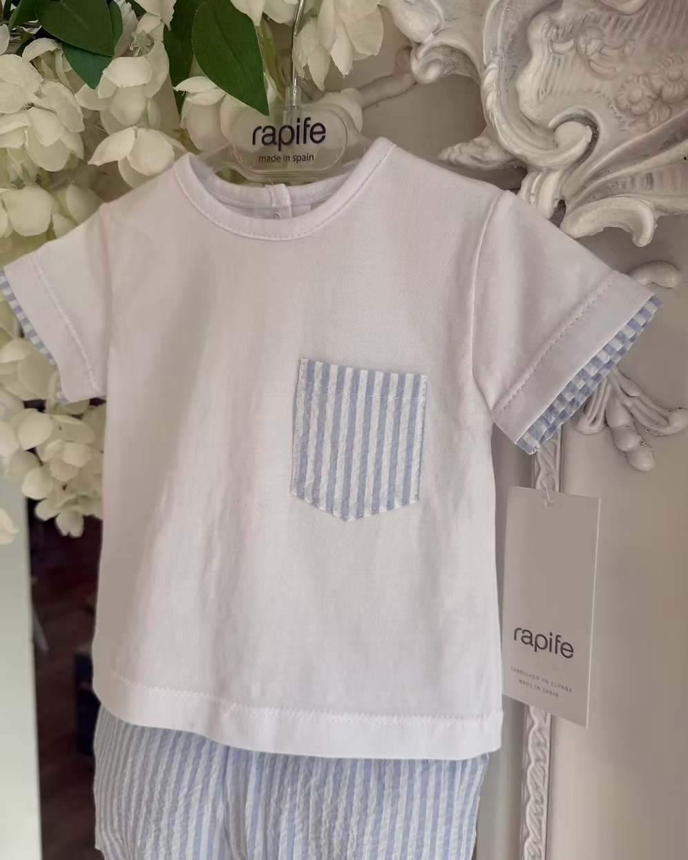 White children's t-shirt with a blue pocket and matching shorts, hanging against a decorative wall.